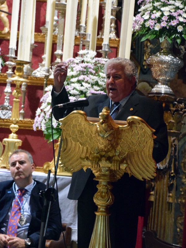 Un hombre habla desde un podio dorado en un entorno festivo con velas y flores.