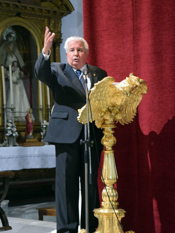 Persona hablando en un púlpito dorado en un entorno religioso con una imagen de la Virgen María en el fondo.