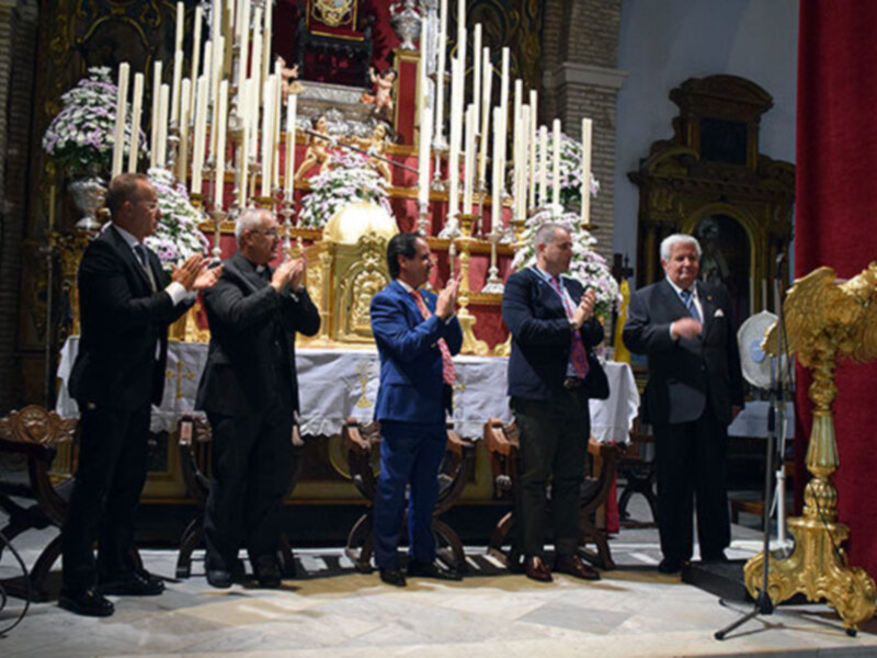 Una ceremonia religiosa en una iglesia, con personas vestidas formalmente aplaudiendo en un altar adornado con flores y velas. La atmósfera es solemnemente festiva.