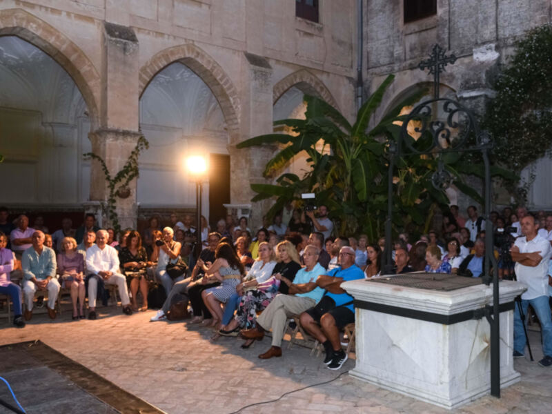 Asistentes a una conferencia o evento en un patio con arcos y palmeras, iluminado por una lámpara de pie.