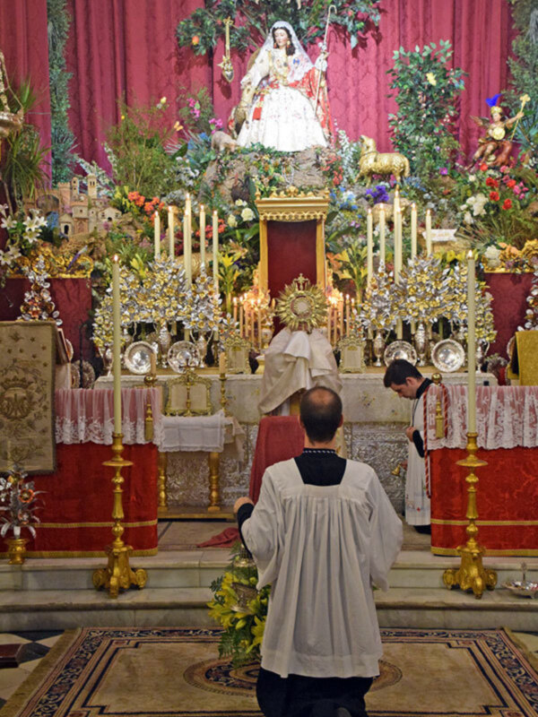 Una imagen de una iglesia con un altar adornado con flores y velas, donde un sacerdote ora ante una imagen de la Virgen María vestida con un vestido blanco y un velo.