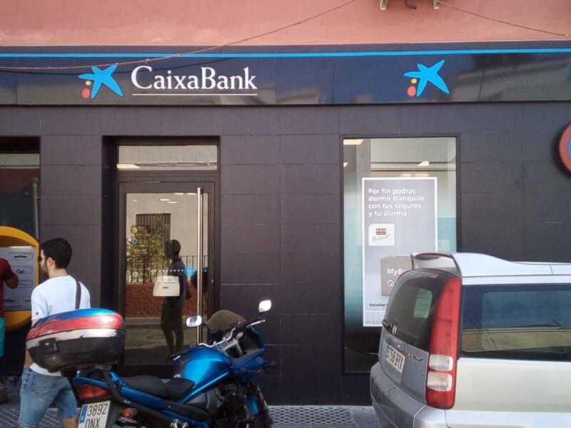 Sucursal de CaixaBank con clientes utilizando cajeros automáticos y un vehículo estacionado frente a la entrada.
