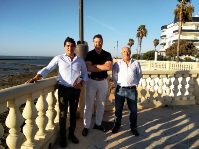 Tres hombres posan juntos en un balcón con vistas al mar y palmeras al fondo.