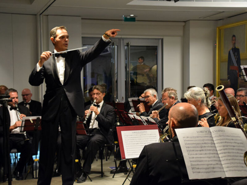 Orquesta dirigida por un maestro en un concierto formal, con músicos tocando instrumentos de viento en un escenario bien iluminado.