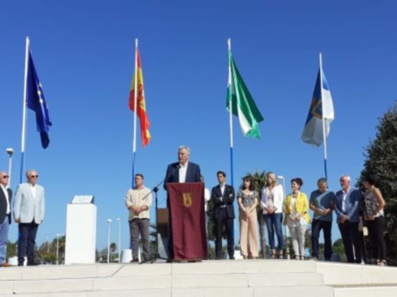 Celebración pública con banderas y discursos en un escenario al aire libre bajo un cielo azul claro.