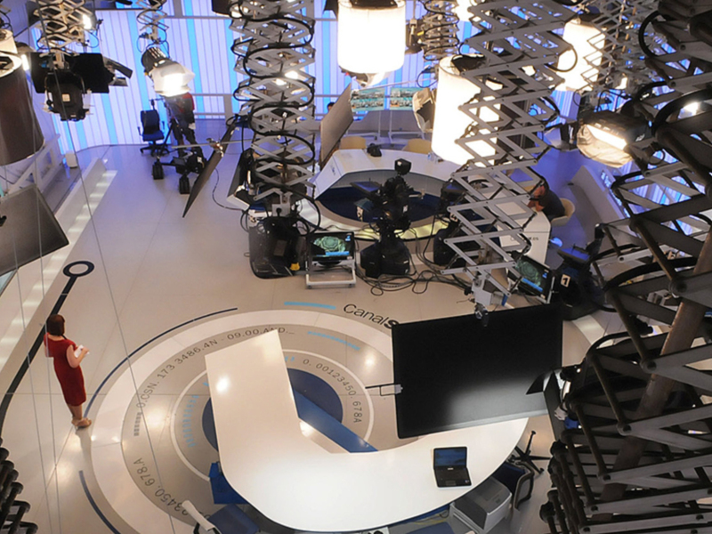 Un estudio de televisión moderno con una mesa redonda en el centro, rodeada de cámaras y monitores. La iluminación es brillante y la decoración es futurista.