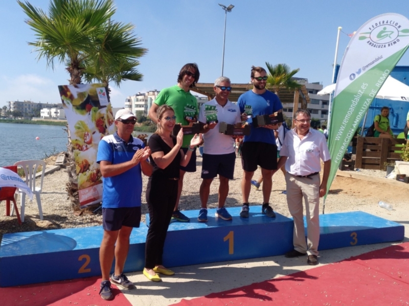 Premiación en el podio de una competencia de remo, con tres ganadores y un representante de la Federación Andaluza de Remo.