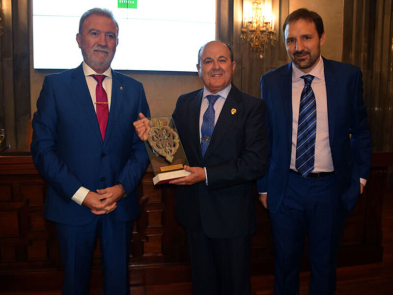 Tres hombres en trajes azules sonríen mientras sostienen un libro con el título "El Cuerpo de la Ley". La imagen se toma en una sala con decoración clásica y luces de araña.