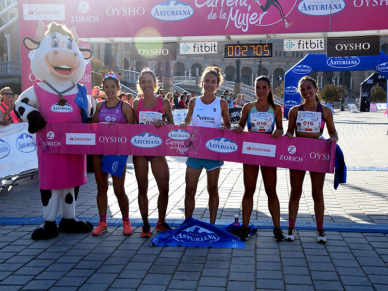 Participantes de una carrera femenina celebrando su victoria en el podio, con una mascota de vaca y una pantalla de tiempo en el fondo.
