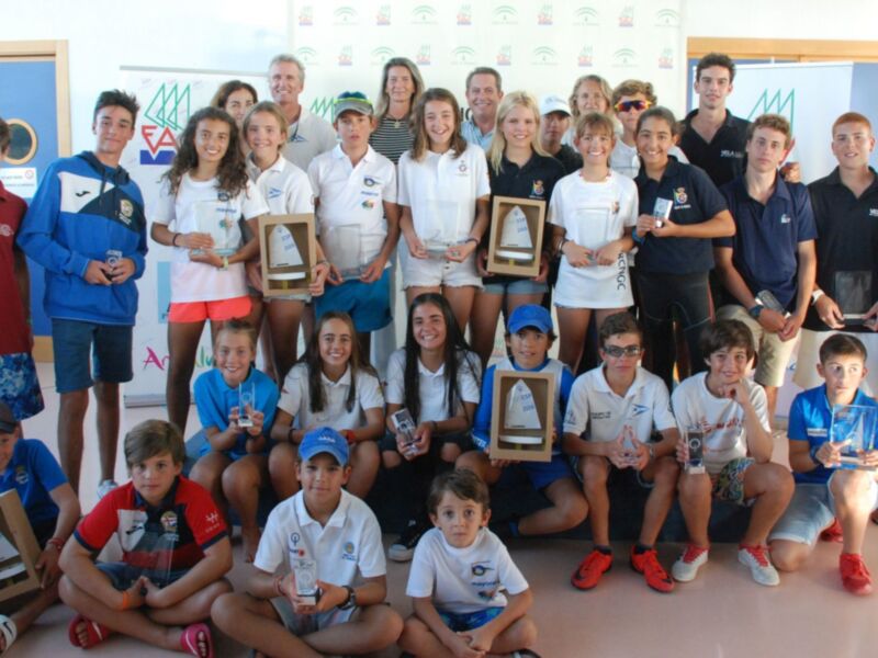 Equipo de jóvenes deportistas posando para una foto de grupo, todos sonrientes y sosteniendo trofeos y medallas, con una pared de fondo con logos de organizaciones deportivas.