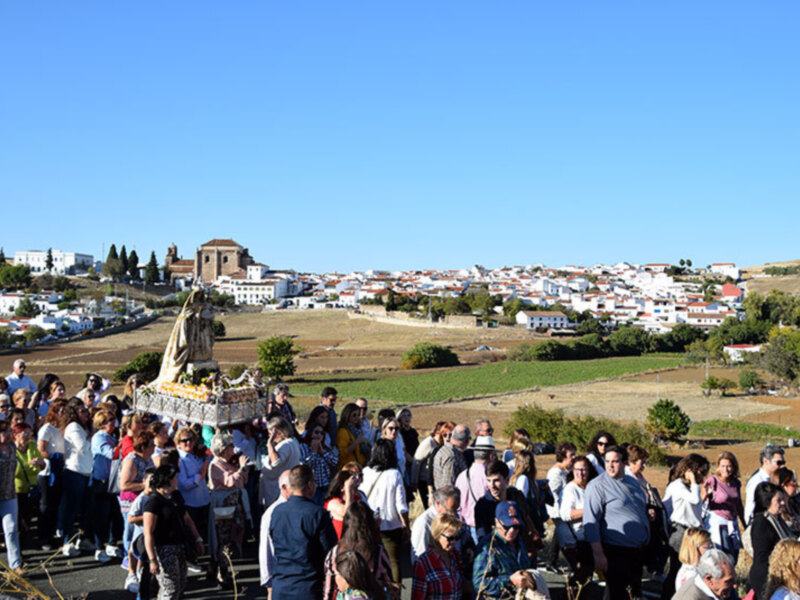 Una multitud de personas participando en una procesión religiosa, con una imagen de la Virgen en el centro, en un paisaje rural con una iglesia en el fondo y un cielo azul claro.