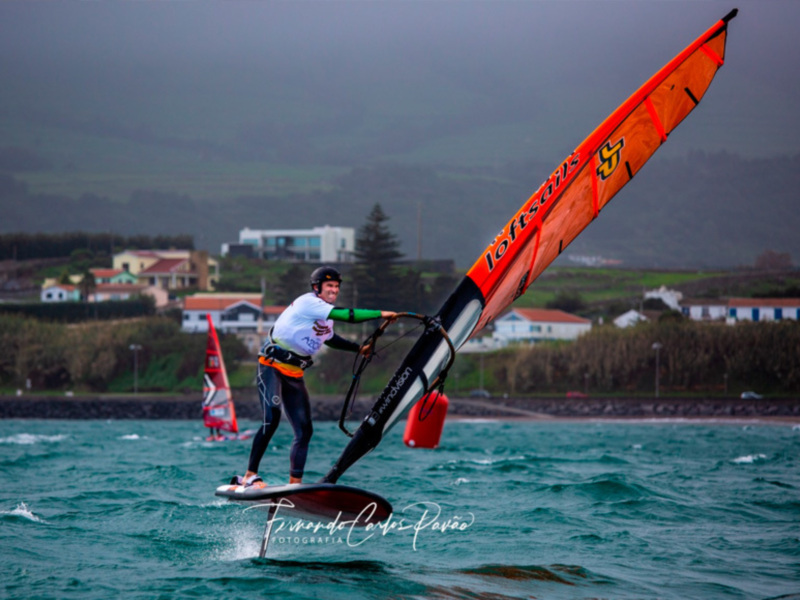 Un windsurfer disfruta de la ola en el mar, con una casa y vegetación en el fondo.