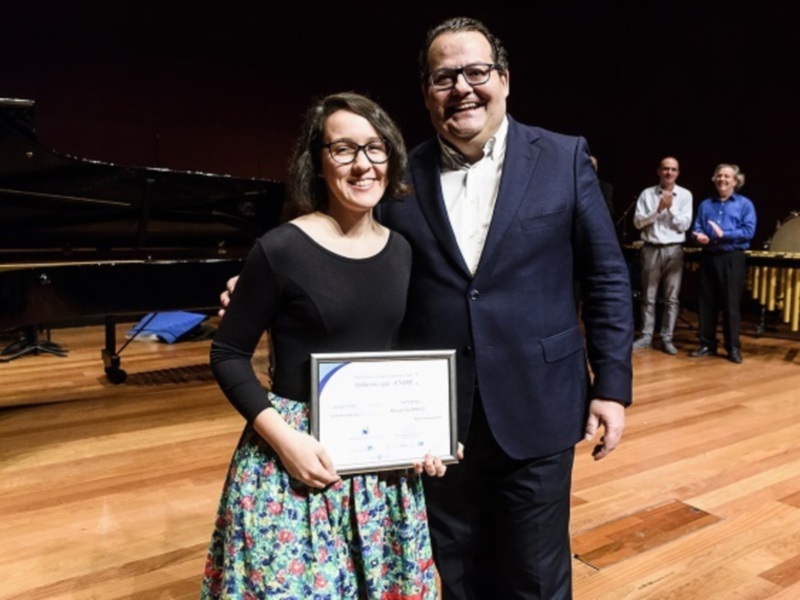 Persona recibiendo un reconocimiento en un escenario musical con piano y tambores en el fondo.