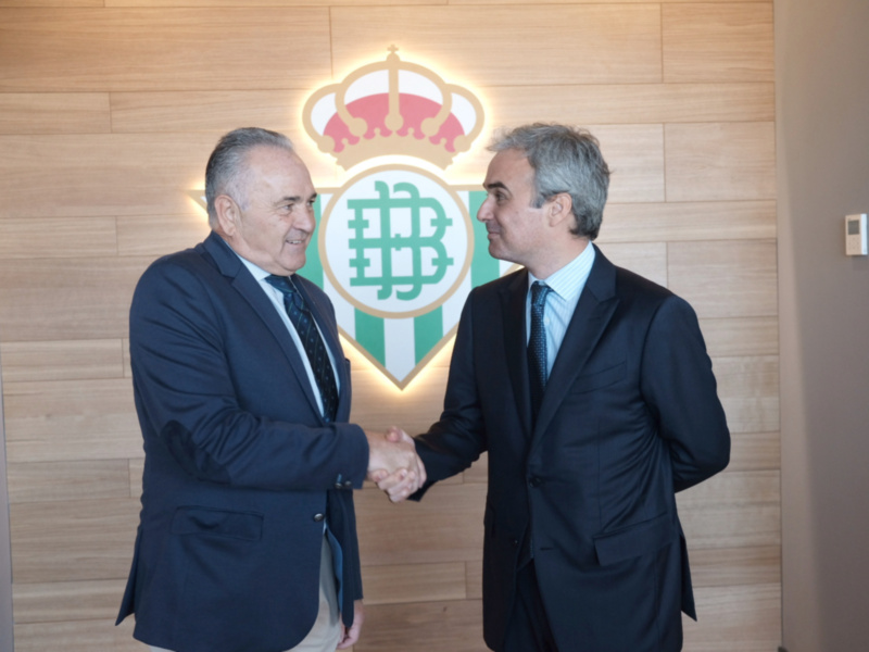 Dos hombres en traje saludándose frente al escudo del Real Betis Balompié.