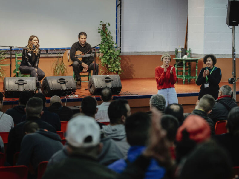 Una mujer en un suéter rojo aplaude mientras una mujer y un hombre tocan guitarra en una escena de concierto en un auditorio.