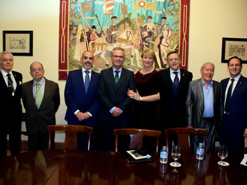 Grupo de personas posando para una foto en una sala formal con una pintura mural en el fondo. Los hombres llevan trajes y los hombres y mujeres llevan camisas y chaquetas. Hay dos vasos de agua en la mesa frente a ellos.