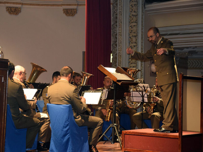 Orquesta militar en un concierto militar, con músicos en uniforme tocando instrumentos de viento y percusión en un escenario con decoración clásica.
