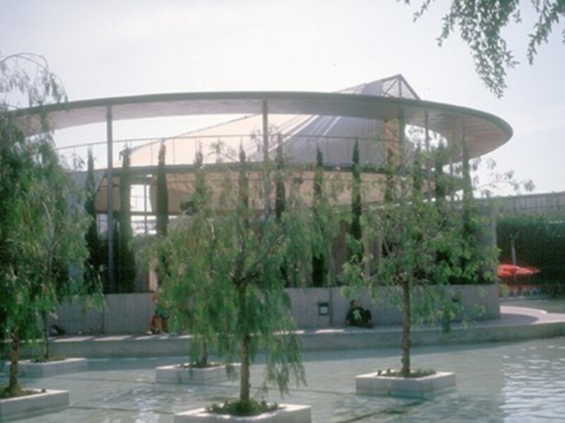 Un pequeño parque con una fuente circular y árboles jóvenes, rodeado por una valla blanca. El cielo es claro y azul, con algunas nubes dispersas. En el fondo, se puede ver una estructura de metal con una cubierta transparente.