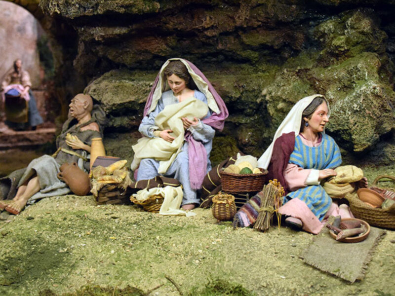 Figuras de madera en un entorno natural, posiblemente una escena de la vida cotidiana de la época medieval, con una mujer sentada con una cesta y una mujer de pie con una bolsa. La escena está ambientada en un entorno rocoso y con vegetación.