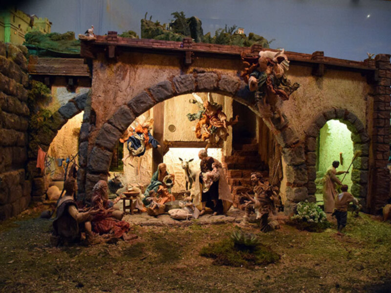 Un belén navideño con escenas de la natividad, incluyendo a los Reyes Magos, un ángel, un burro y un oveja, en una escena con una arquitectura medieval.