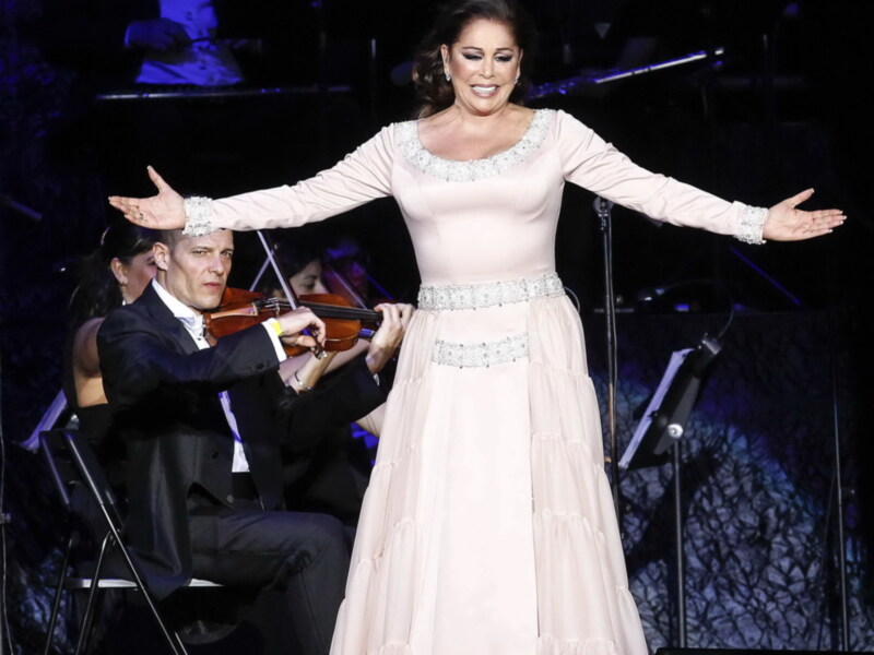 Una cantante en un escenario con orquesta, vestida con un vestido largo de color rosa claro, con el brazo extendido en un gesto de presentación o celebración.