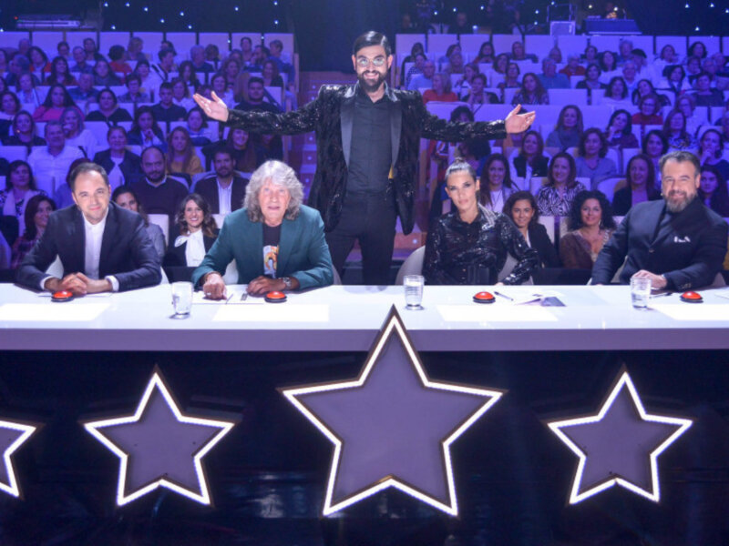 Júri en un escenario de talentos con estrellas luminosas en primer plano y audiencia en el fondo.
