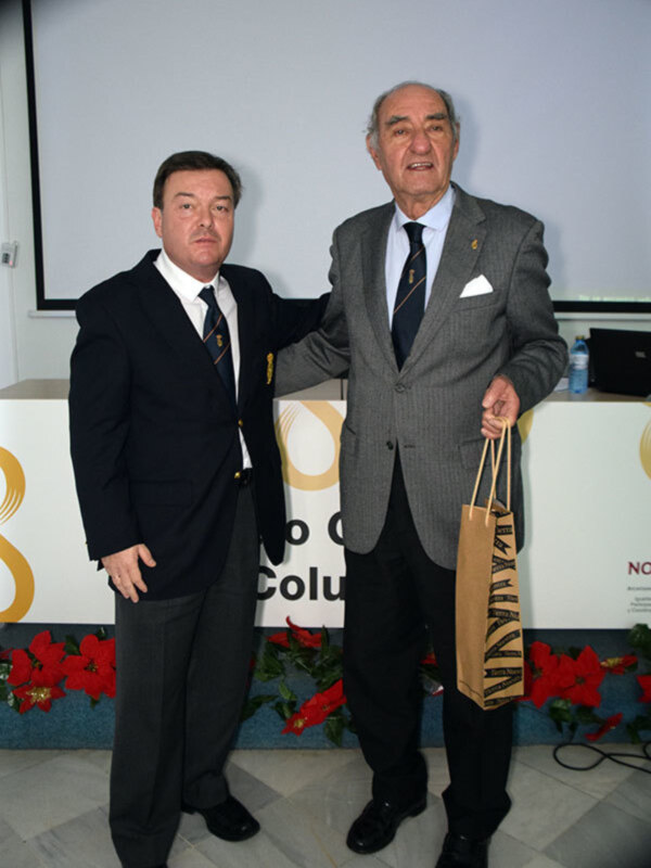 Dos hombres en trajes posan juntos en un evento formal, uno sosteniendo un bolso de regalo.
