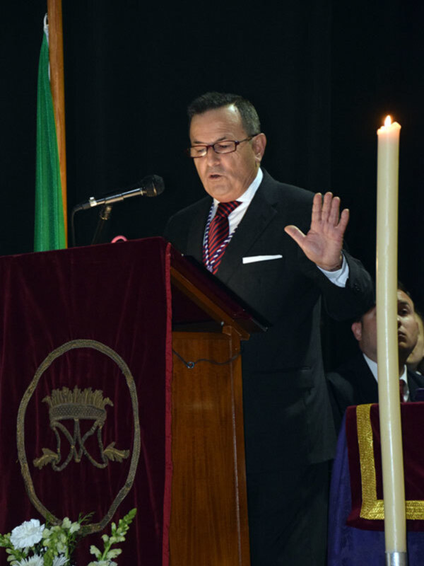 Un hombre en traje habla desde un podio durante una ceremonia formal, con una vela encendida y una bandera verde detrás.