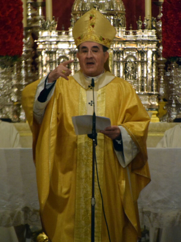 Un sacerdote vestido con un manto amarillo y un hábito de color dorado, con una cruz en el pecho, dirigiendo una oración en una iglesia con una gran cúpula dorada y velas rojas en el fondo.
