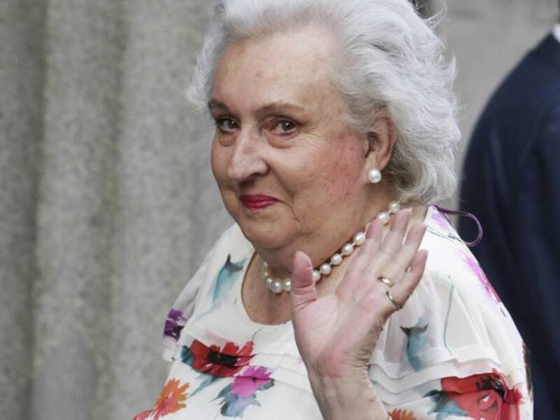 Retrato de una mujer mayor con flores y perlas, saludando con la mano derecha.