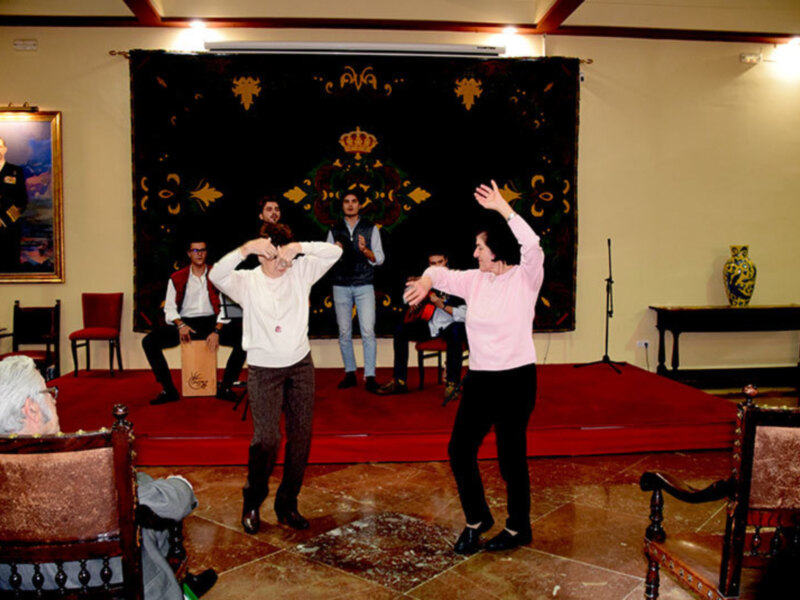 Actores en escena durante una representación teatral en un salón decorado con tapiz y sillas antiguas.