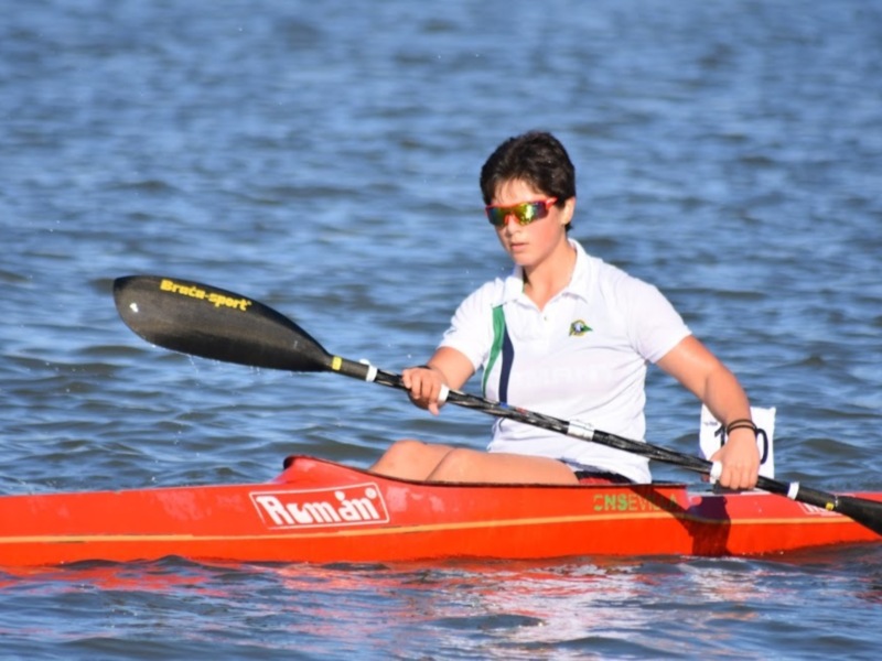 Un joven kayakista en acción, con una camiseta blanca y gafas de sol, remando en un kayak rojo en aguas tranquilas.