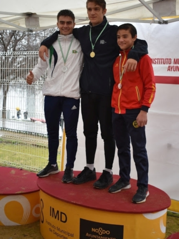 Tres atletas posan con sus medallas en un podio, con una valla y un edificio en el fondo.