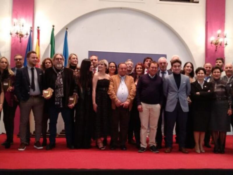 Una celebración formal con personas posando para una foto en un escenario rojo, con banderas y luces en el fondo.