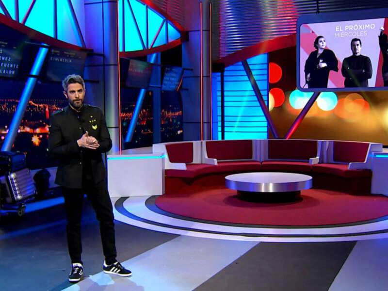Un presentador en un estudio de televisión moderno, con un fondo de luces y un televisor en la pared que muestra una imagen de dos personas.