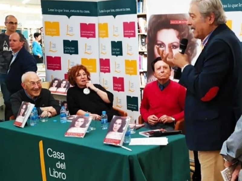 Presentación de libros en una librería, con autor y público en una mesa con carteles promocionales.