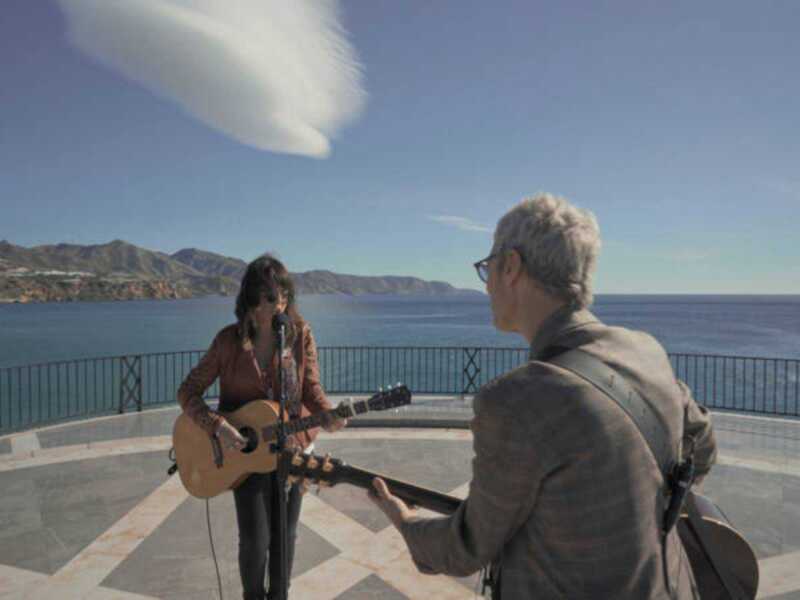 Dos músicos tocan guitarra en un balcón con vista al mar y montañas.