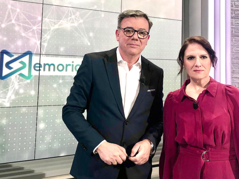 Dos presentadores en un estudio de televisión, con un fondo que muestra un diseño de red digital y la palabra 'Memoria'.