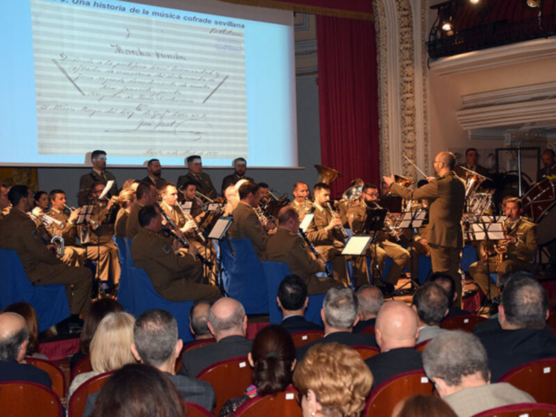 Concierto de banda militar con proyección de partituras históricas en un escenario teatral.