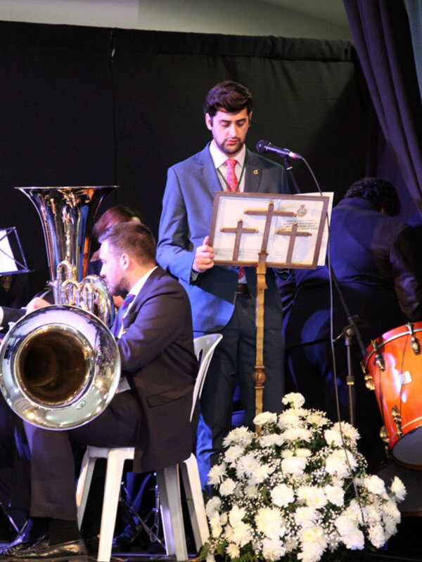 Un hombre en traje sostiene un marco con un diploma mientras otros músicos tocan en el fondo. Hay una gran florero de flores blancas en el frente de la imagen.