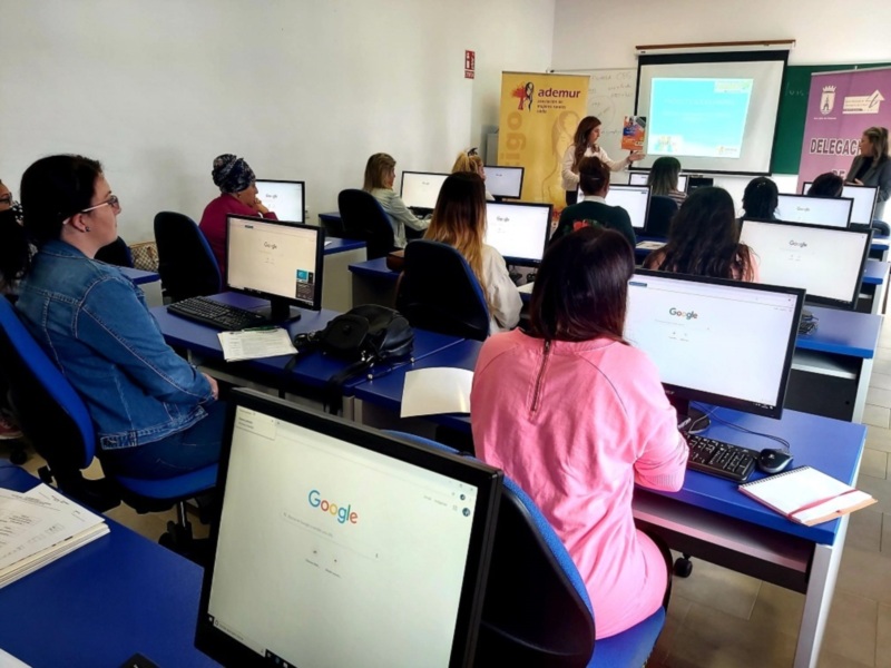 Clase de informática con estudiantes trabajando en computadoras y una profesora explicando en la pizarra.