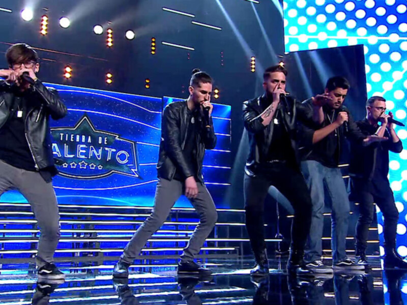 Un grupo de cinco personas en una escena de concierto, con luces y un fondo azul con estrellas. Los individuos están vestidos con ropa casual y están cantando en micrófonos. La escena tiene un ambiente de espectáculo y entretenimiento.