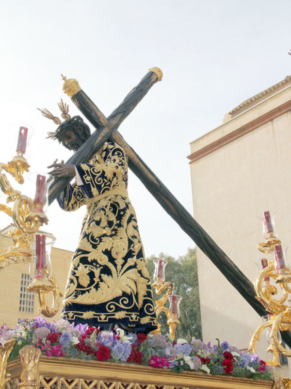 Estatua de Cristo con cruz en procesión, rodeada de flores y velas, con un edificio en el fondo.