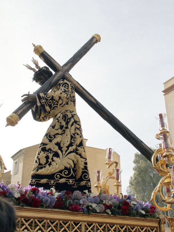 Procesión de Semana Santa con imagen de Jesús portando la cruz, vestido con traje negro y dorado, en un carro ornado con flores y candelabros dorados.
