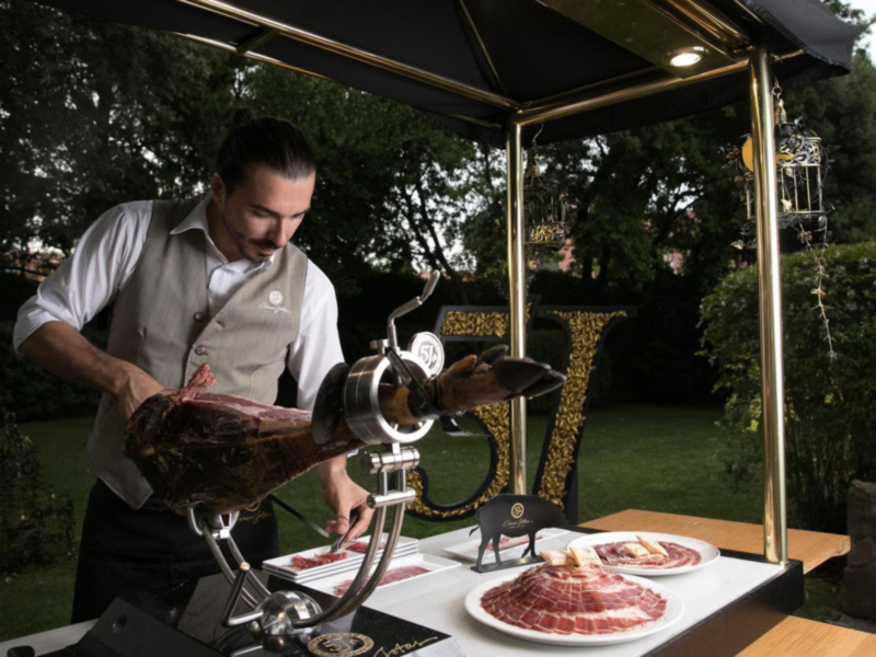 Un chef prepara un asado de carne de res en un mostrador de carnes, iluminado por una lámpara dorada, en un entorno exterior con vegetación y un mueble de madera dorada en el fondo.