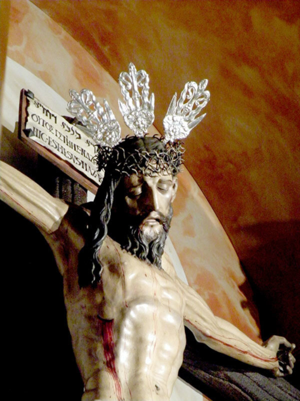 Escultura de Cristo crucificado con detalles de la corona de espinas y la sangre en la mano derecha, en un contexto religioso y artístico.