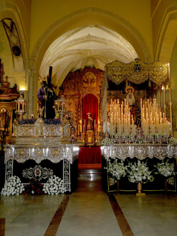 Interior de una iglesia con altares ornamentados y candelabros de velas, decoración religiosa y flores blancas.