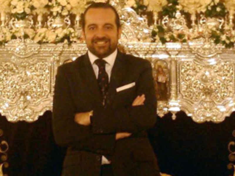 Un hombre en traje negro con las manos cruzadas frente a una mesa decorada con flores y adornos.