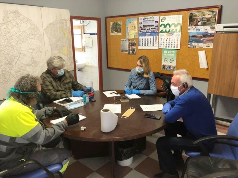Reunión de trabajo en un entorno de oficina, con participantes usando mascarillas y guantes, en un ambiente de discusión y colaboración.