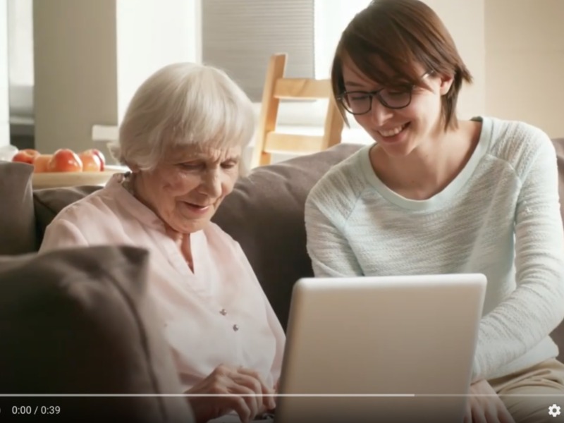 Una joven y una anciana sonríen mientras trabajan en un laptop en un ambiente acogedor. La escena transmite un sentimiento de apoyo y aprendizaje mutuo.